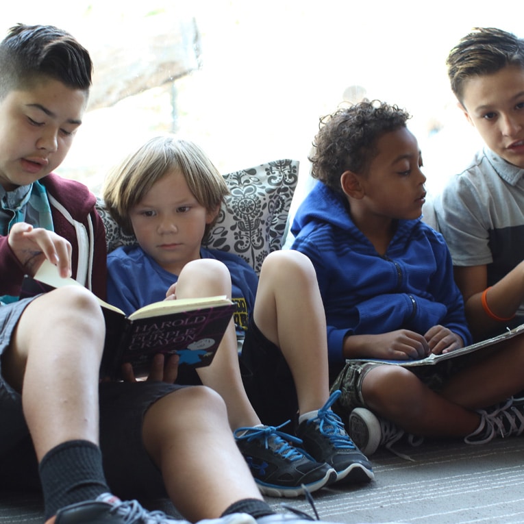 Four boys sit closely together on the floor, reading books. They appear engaged, with two boys looking at the same book while the others focus on books in their laps. Cushions and soft light create a cozy atmosphere.