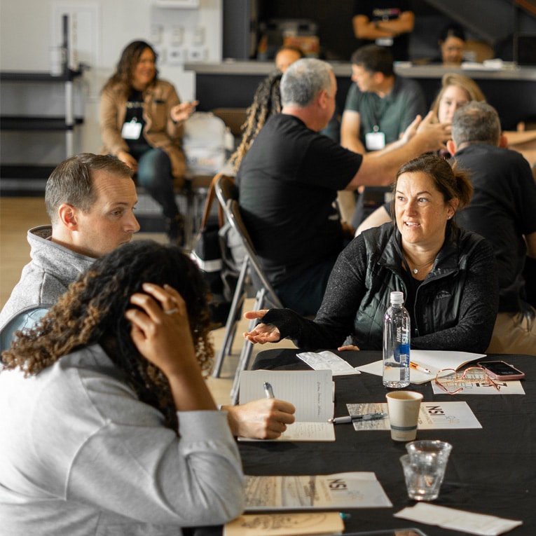 A group of people sit around tables in a bright room, engaged in discussion and taking notes. Some are talking, while others listen or write. Papers, cups, and a water bottle are on the table in the foreground.