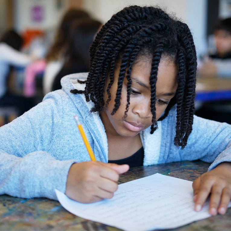 A young girl with braided hair sits at a desk, focused on writing with a pencil on a sheet of paper in a classroom setting. Other students are blurred in the background.