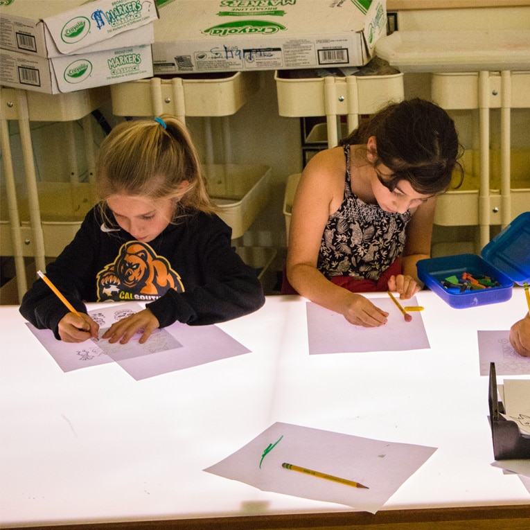 Two young girls sit at a brightly lit table, drawing on white paper with pencils. Art supplies and boxes are visible in the background.