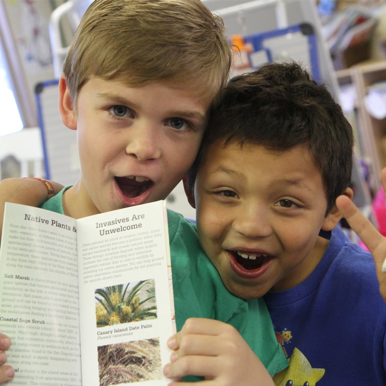 Two young boys smile and pose together while holding an open pamphlet about invasive plants. The boy on the left has his arm around the other, and both look excited and engaged.