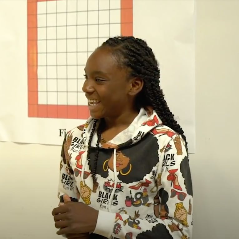 A smiling girl with braided hair stands in front of a wall chart. She is wearing a white hoodie with colorful graphics and the words BLACK GIRLS RUN! printed on it.
