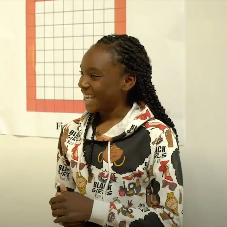 A smiling girl with braided hair stands in front of a wall chart. She is wearing a white hoodie with colorful graphics and the words BLACK GIRLS RUN! printed on it.