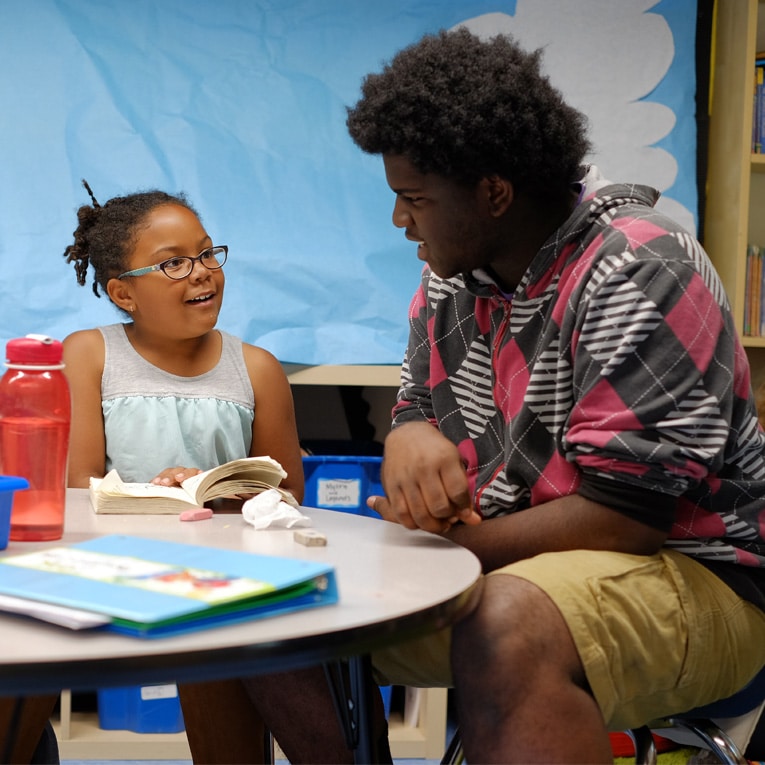 A young girl smiles while reading a book at a table with an adult who listens attentively. They appear to be having an engaging conversation in a classroom setting.