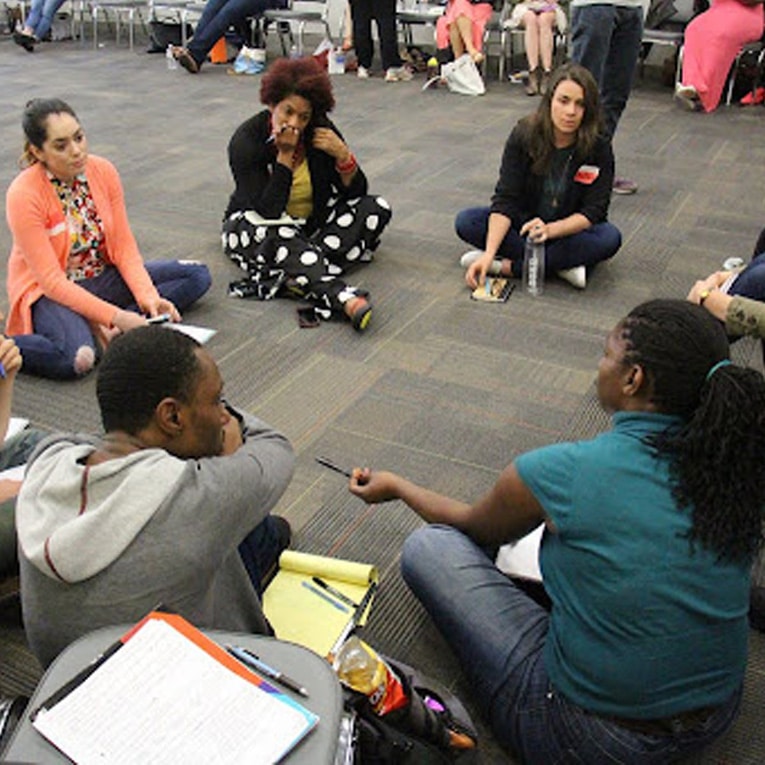 A diverse group of people sits in a circle on a carpeted floor, engaged in discussion. Notebooks, pens, and water bottles are scattered around as they listen and interact in a casual, collaborative setting.