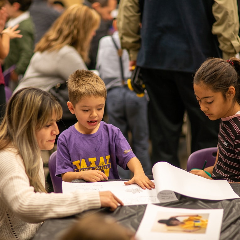A young boy in a purple shirt sits at a table with a woman and a girl, looking at papers and drawing in a busy classroom or event setting.