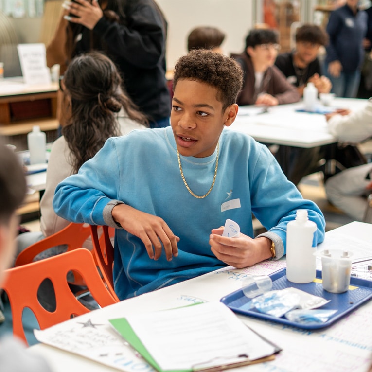 A teenage boy in a blue sweatshirt sits at a table with science materials, talking to others in a busy classroom filled with students working on projects.
