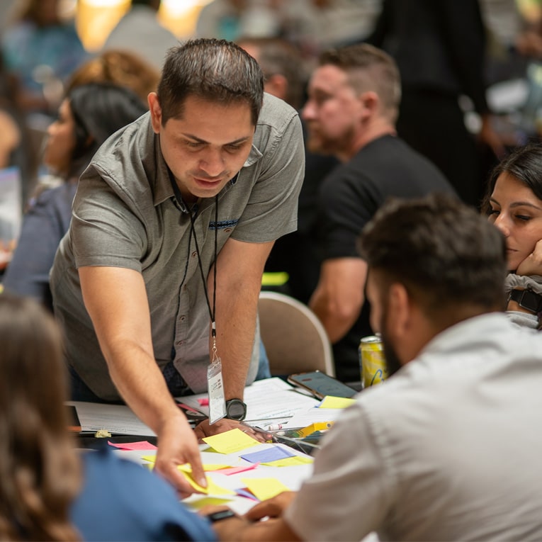 A man leans over a table covered with colorful sticky notes, engaging with others in a collaborative group discussion. People are seated around him, focused on the activity. The setting appears to be a workshop or meeting.
