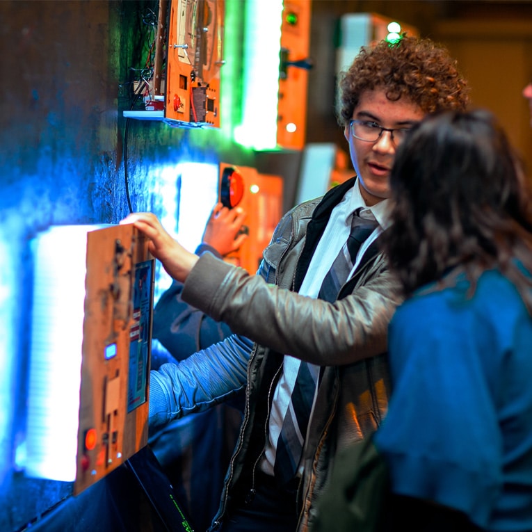 A young man in a suit and jacket interacts with an illuminated wall display at an event, talking to a woman beside him. The background glows with colorful lights and other people are also engaging with the exhibits.