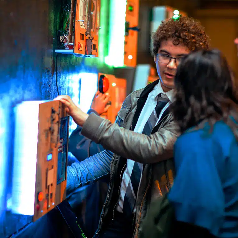 A young man in a suit and jacket interacts with an illuminated wall display at an event, talking to a woman beside him. The background glows with colorful lights and other people are also engaging with the exhibits.