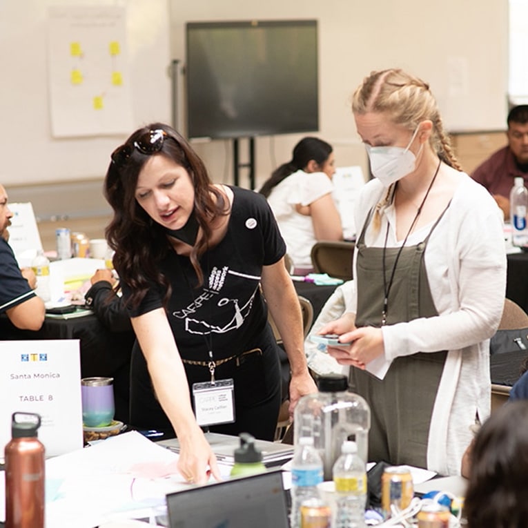 Two women collaborate at a busy conference table, discussing a planned experiment. One points to some papers while the other, wearing a face mask, listens. Other attendees focused on improvement work in the bright, indoor setting.