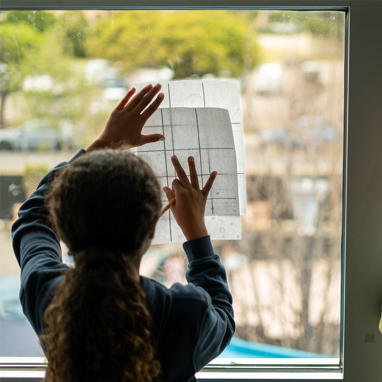 A child with curly hair holds a sheet of paper with a grid against a window, tracing or drawing while looking outside. The scene outside the window is blurred with trees and parked cars visible.