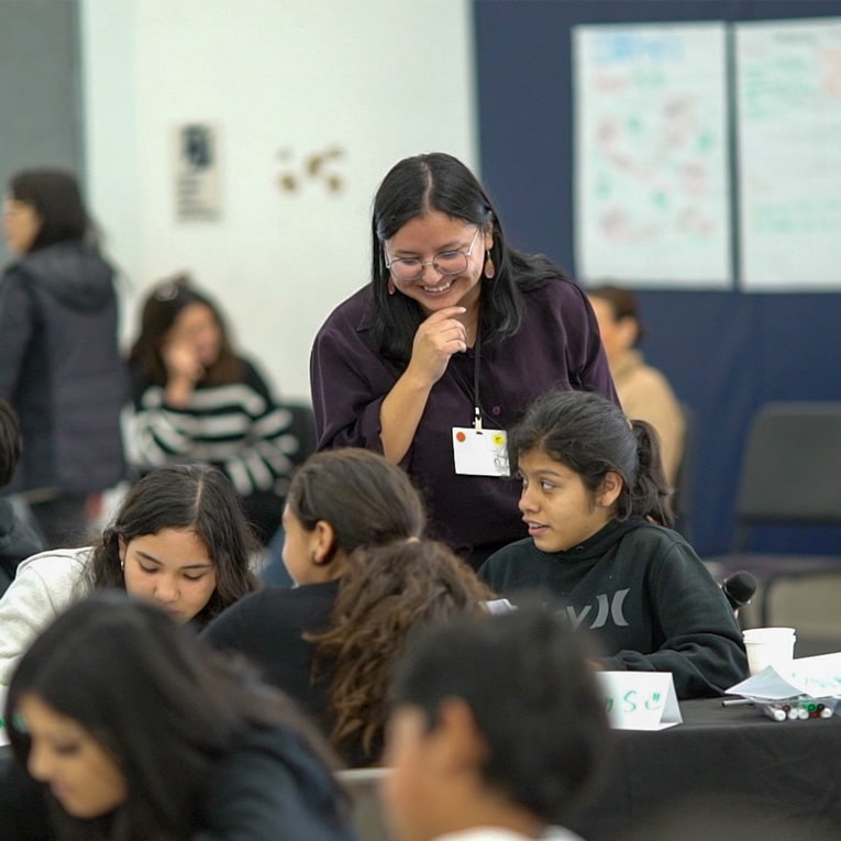 A smiling woman with glasses leans over a table, engaging with a group of young students who are working together in a classroom setting. Other people and educational materials are visible in the background.