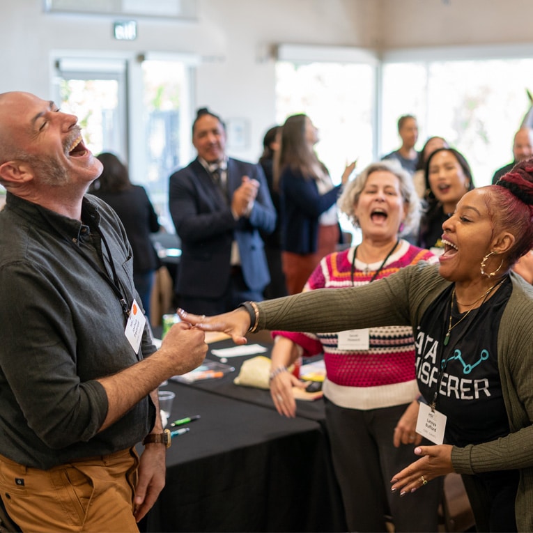 A group of people indoors laughing and interacting enthusiastically, with two individuals in the foreground reaching toward each other, surrounded by others clapping and smiling during a lively event or workshop.