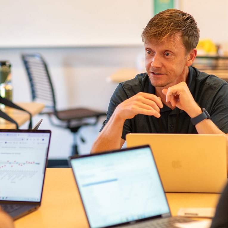 A man with light brown hair sits at a table, engaged in conversation during a meeting. Two open laptops with charts on their screens are in the foreground. The background shows an office setting with chairs and a blurred whiteboard.