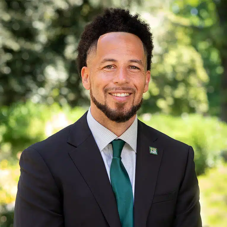 A man with short curly hair and a beard, wearing a dark suit, white shirt, and green tie, smiles while seated outdoors with greenery and trees blurred in the background.