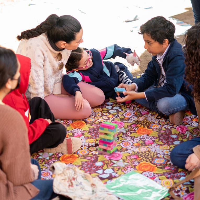 A group of children and adults sit on a colorful floral blanket outdoors, joyfully playing a game with stacked, multicolored blocks—smiling together as they interact and subtly swim against the current of white dominant culture.
