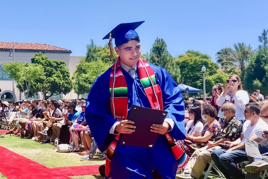 A graduate in a blue cap and gown holds a diploma while walking outdoors at a graduation ceremony, celebrating achievement in Priority Areas. The audience sits on either side, with trees and buildings under a clear sky in the background.