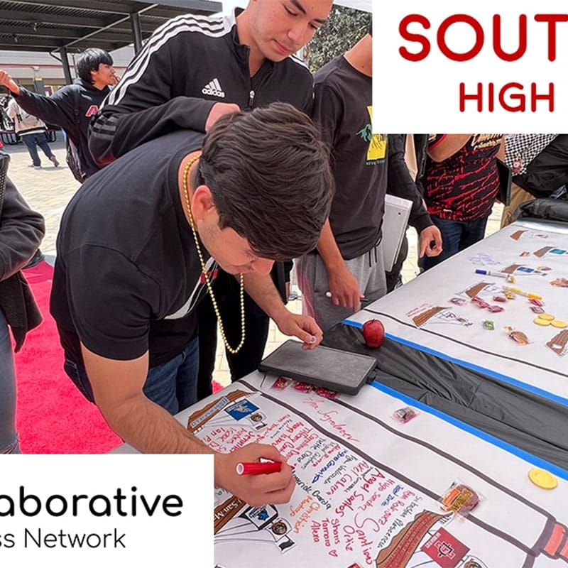 A student writes on a large poster at a table during an outdoor school event at Southwest High School. Other students stand nearby, participating in the activity. Parts of signs read SOUTH HIGH and Collaborative Business Network, highlighting this moment as a Story of Impact within the school community.