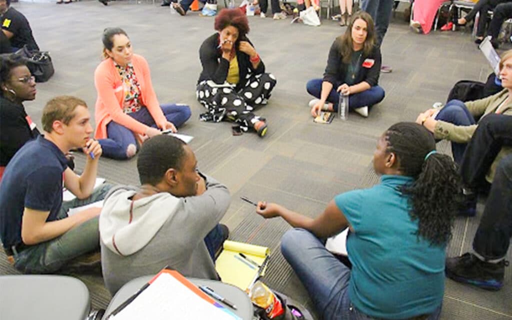 A diverse group of people sit in a circle on the floor, engaged in a discussion about resources for continuous improvement. Some hold notebooks and pens, suggesting a collaborative or workshop setting. The background shows more people seated similarly.