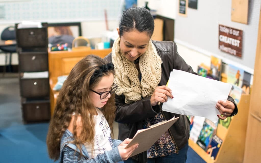 A young girl and an adult woman stand together in a classroom, looking at papers. The girl holds a clipboard while the woman, wearing a scarf and jacket, smiles and explains something about resources for continuous improvement. Books line the background.