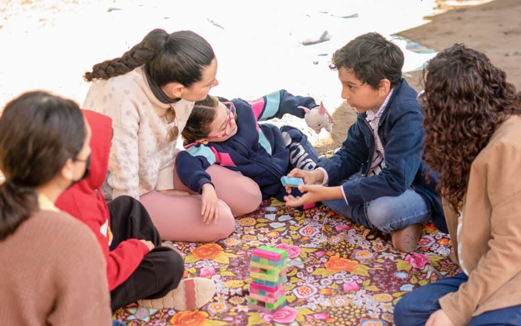 Five children sit together on a colorful patterned blanket outdoors, playing Jenga and talking, as they use teamwork and shared resources to enjoy continuous improvement in their game and friendship.