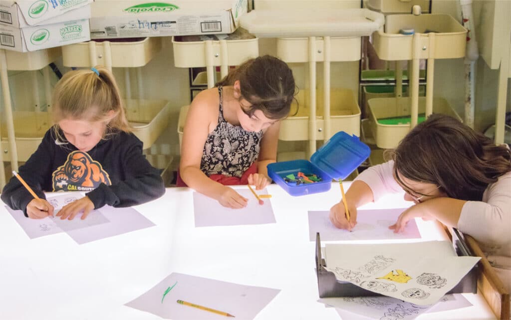 Three young children sit at a lit table, focused on drawing with pencils. Colored pencils, sketches, and continuous improvement resources are scattered on the table, while storage carts and other supplies are visible in the background.