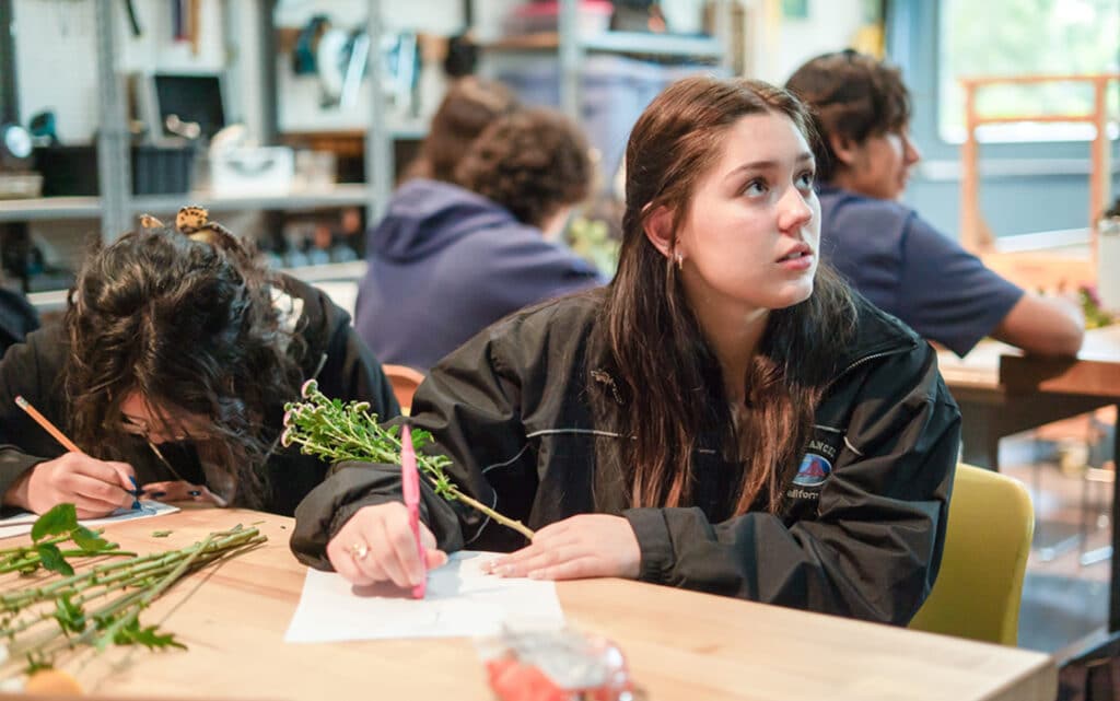 A young woman in a black jacket sits at a table in a classroom, holding a pen and a stem with leaves, looking up attentively as she explores improvement resources. Other students nearby write and work with plants.