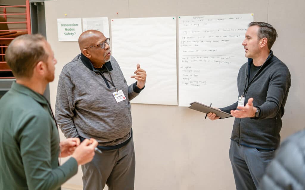 Three men stand before a wall of blank and handwritten papers, discussing resources for continuous improvement. One holds a tablet, and all wear conference badges, highlighting a collaborative work or meeting environment.