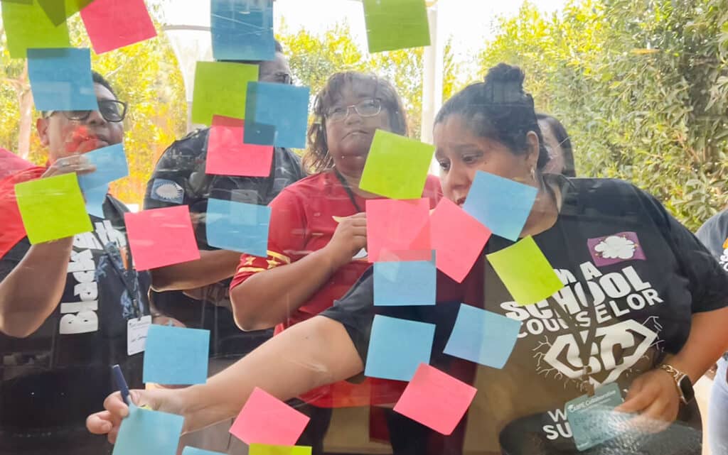 A group of people stand behind a glass wall covered in colorful sticky notes. One woman writes on a note while others look on attentively, using Continuous Improvement Resources to enhance their collaborative activity.