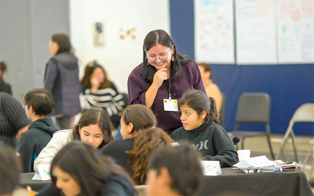 A smiling woman stands and interacts with a group of students working together at a table in a classroom, fostering collaboration and sharing continuous improvement resources. Other students and adults are visible in the background.