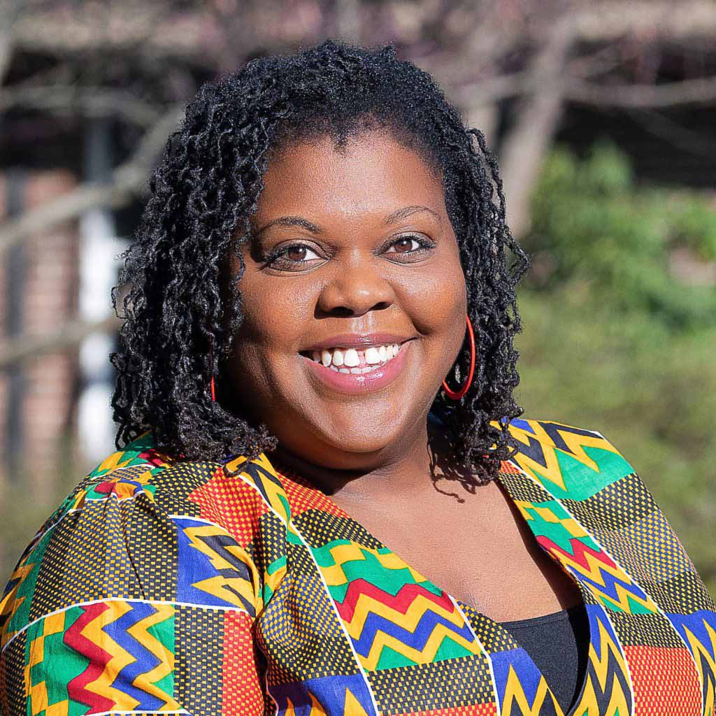 A woman with curly black hair and hoop earrings smiles outdoors, wearing a colorful patterned top with yellow, green, red, and black geometric designs—reminiscent of Brandi Hinnant-Crawford’s vibrant style.