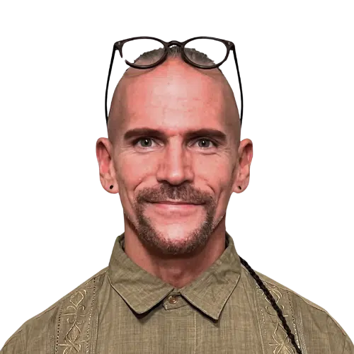A smiling bald man, identified as Derek Mitchell, with a trimmed mustache and goatee wears glasses on his head, a buttoned olive green shirt, and small earrings. The background is transparent.