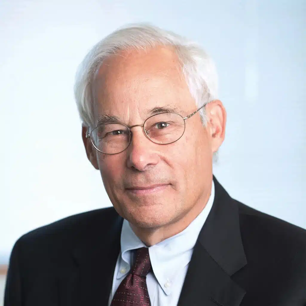 An older man with white hair and glasses, wearing a dark suit, light blue shirt, and patterned tie, poses in front of a light, blurred background.