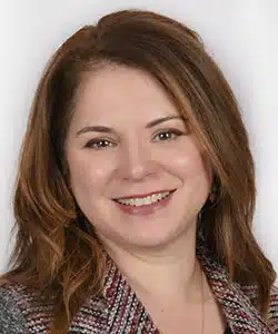 A woman with shoulder-length brown hair smiles at the camera. She is wearing a patterned jacket and a light-colored top, posing against a neutral background for Derek Mitchell’s latest portrait series.