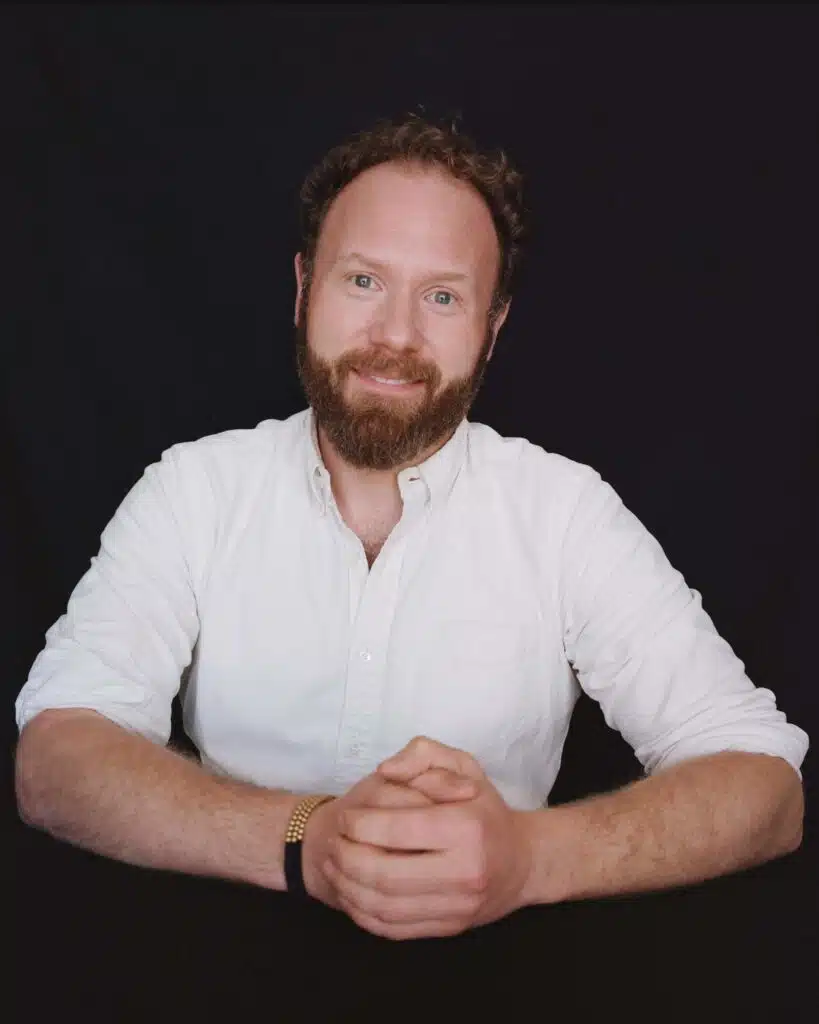 A man with curly hair and a beard, identified as Justin Cohen, wearing a white button-up shirt, sits at a table with his hands clasped and smiles at the camera against a black background.