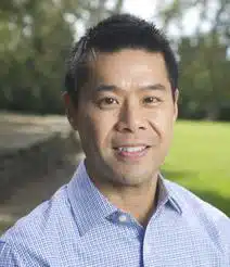 A man with short dark hair, wearing a light blue checked shirt, smiles outdoors. The background shows greenery and trees in soft focus.