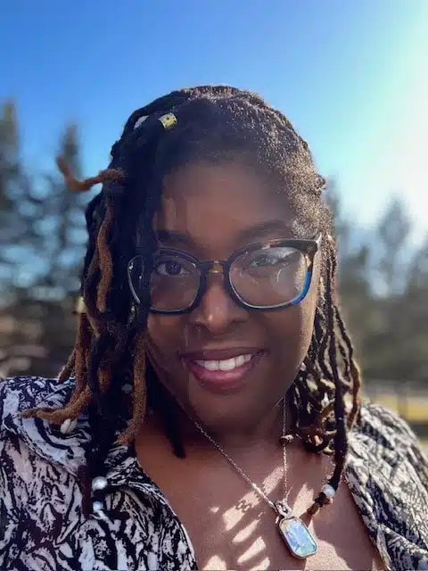 Smiling Nichelle Woodson, with glasses and long, beaded dreadlocks, stands outdoors on a sunny day, wearing a patterned shirt and a pendant necklace. Trees and a bright blue sky are visible in the background.