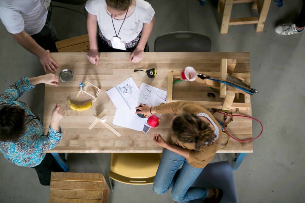 Three people gather around a wooden table covered with papers, sketches, tools, a banana, and small models, collaborating on a hands-on design project—capturing the spirit of learning at the PBL Leadership Academy.