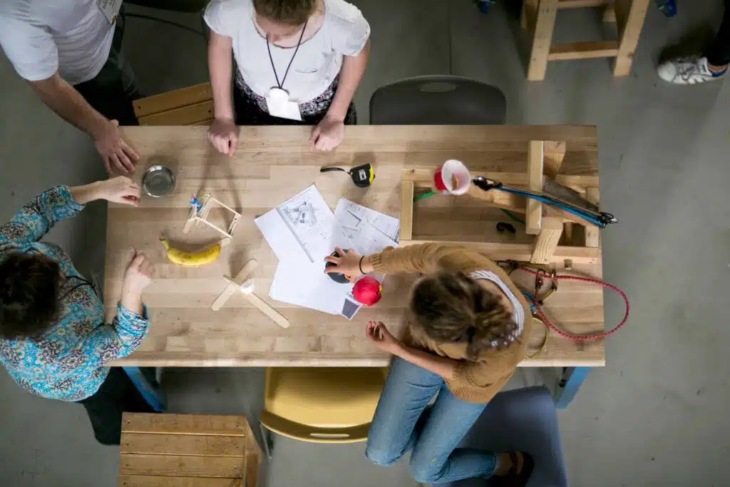 Three people gather around a wooden table covered with papers, sketches, tools, a banana, and small models, collaborating on a hands-on design project—capturing the spirit of learning at the PBL Leadership Academy.