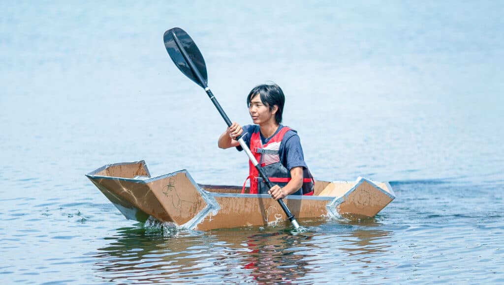 A person paddles a makeshift boat made of cardboard on a calm body of water, wearing a life jacket and holding a black paddle—a creative scene reminiscent of Project Unpacking ingenuity.