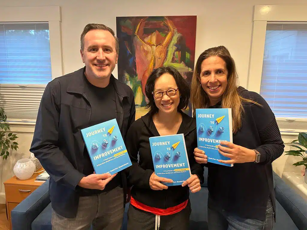 Three people—Sandra Park, Alicia Grunow, and Brandon Bennett—smiling and holding copies of the book Journey to Improvement in a cozy living room with art, plants, and a window in the background.