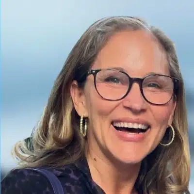 A woman with shoulder-length light brown hair, wearing glasses, large hoop earrings, and a dark patterned top, smiles broadly against a blurred blue background at Derek Mitchell’s event.