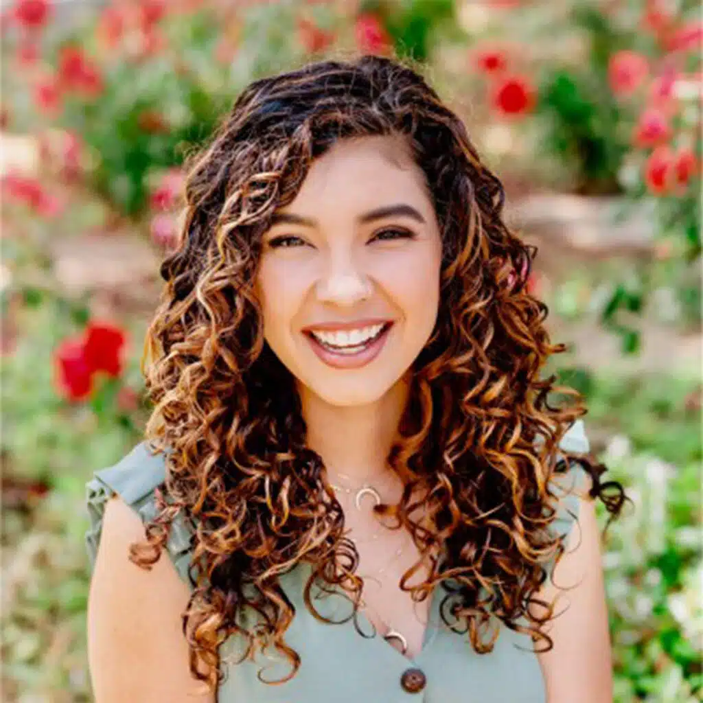 Smiling woman, Vanessa Ruiz, with curly brown hair wearing a light green top, standing outside with red and green plants blurred in the background.