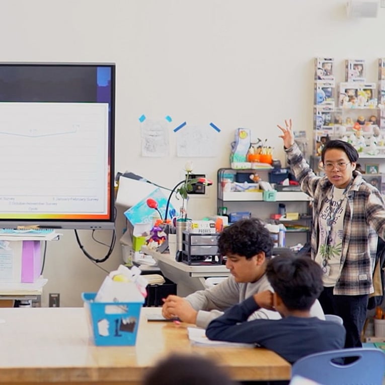 A person stands at the front of a classroom gesturing while presenting to students seated at a table. A large screen displays a graph, highlighting data sharing, and the room is filled with colorful supplies and decorations.