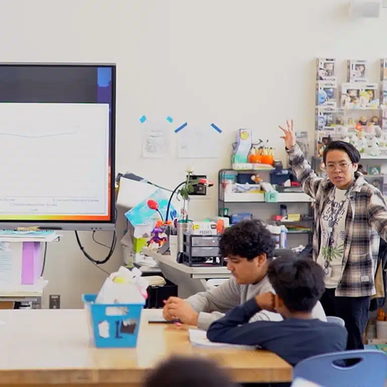 A person stands at the front of a classroom gesturing while presenting to students seated at a table. A large screen displays a graph, highlighting data sharing, and the room is filled with colorful supplies and decorations.