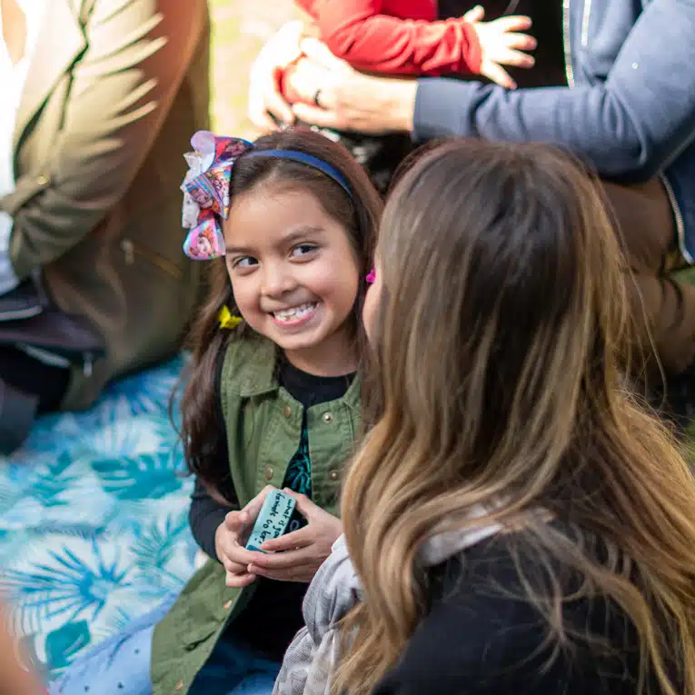 A young girl with a colorful bow smiles, holding a small object, while sitting outdoors on a patterned blanket among other people—a moment that beautifully reflects empathy in design, where thoughtful details create spaces for connection and joy.