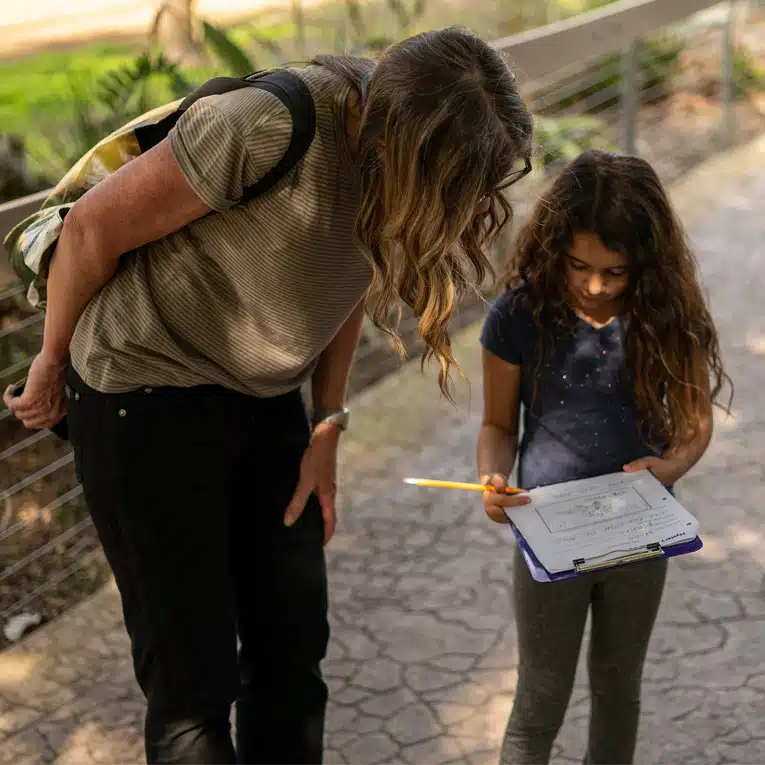 An adult leans over to look at a worksheet held by a young girl with long hair, who is writing on a clipboard outside on a paved path, demonstrating how empathy in design can be fostered through collaborative learning and thoughtful engagement.