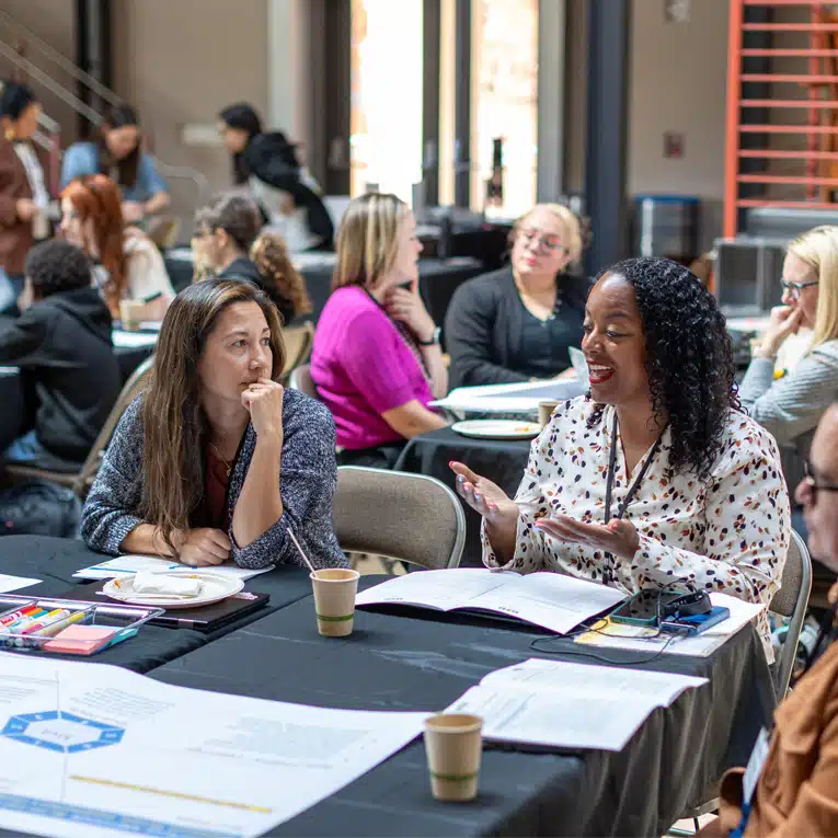 People sit at tables in a bright room, engaged in discussions. Two women in the foreground talk and smile, surrounded by papers, notebooks, Work Books, and coffee cups as they focus on evaluating their Aim Statements. Other groups are visible in the background.