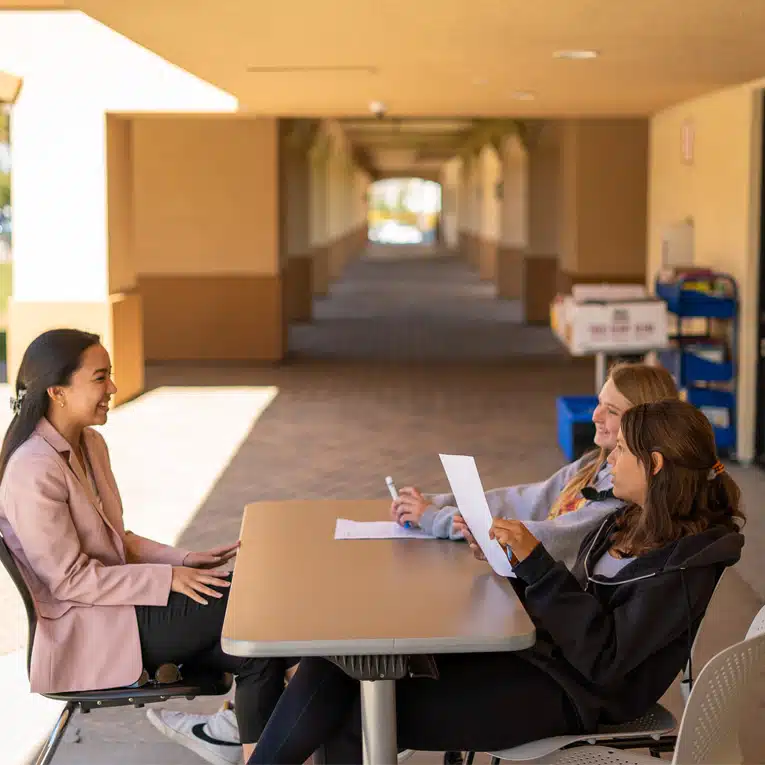 Three people sit at a table outdoors under a covered walkway. Two interviewers face a candidate who is smiling, embodying the spirit of professional development. The background shows the long corridor and some carts with supplies along the wall.